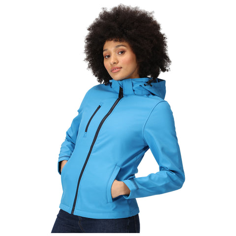 A woman with curly hair poses casually in a bright blue jacket with a hood her hands are in the pockets and she is set against a plain white background