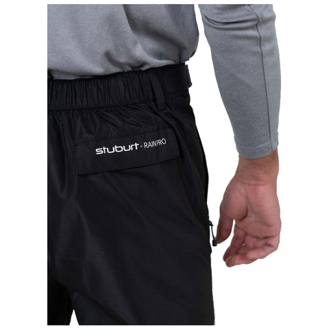 A person's hand reaches towards the waist of black rain pants with a logo label reading "stuburt RAINPRO" visible against a gray shirt in a neutral setting.