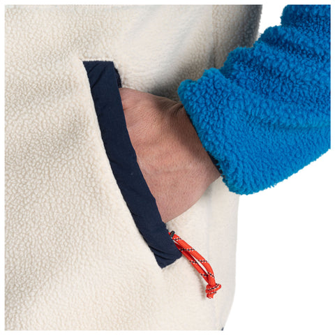 A hand rests inside a pocket of a cream-colored fleece garment while wearing a bright blue fleece sleeve with a textured surface in a neutral setting.