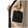 A hand grips an unzipped jacket revealing a plush, beige lining and a small black pocket sewn on the interior, set against a plain backdrop.