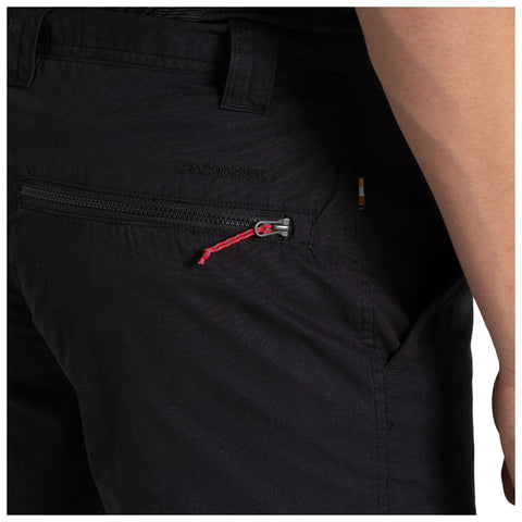 A pair of black hiking pants is displayed with a side view highlighting a zipper pocket that has a red pull tab situated just above the waistband and features a brand logo embossed on the fabric.