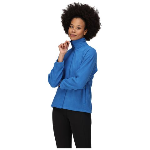 A woman wearing a blue fleece jacket poses with one hand on her chin while standing against a plain white background, presenting a thoughtful expression.