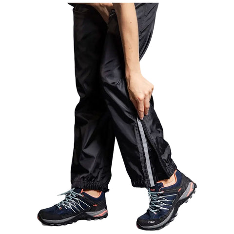 A person is partially unzipping black waterproof pants while wearing sports shoes with blue laces, standing possibly outdoors or in a casual setting with a focus on outdoor activity.