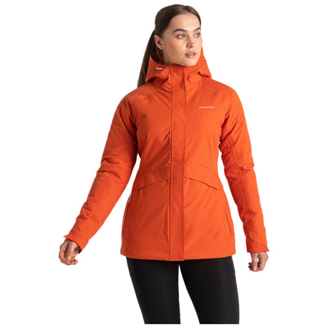 A woman wears a bright orange outdoor jacket with a hood holding her left hand at her waist in a neutral setting, suggesting readiness for outdoor activities.