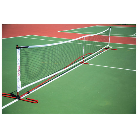 A tennis net stands upright on a green court with red tennis courts visible in the background under bright lighting showcasing a well-maintained sports environment