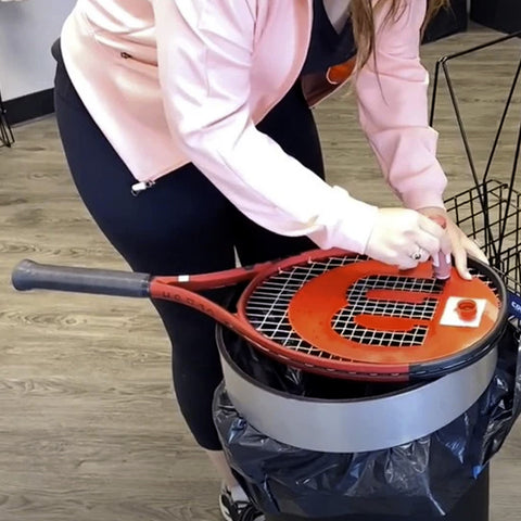 A tennis racket is positioned on top of a trash can while a person applies paint with a brush to the racket's strings in a casual indoor setting with wooden flooring.