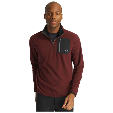 A man stands wearing a burgundy fleece pullover with a half-zip and a black pocket featuring the initials CK, in a neutral setting.