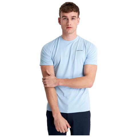 A young man stands with his arms crossed wearing a light blue short-sleeved shirt while looking directly at the camera against a plain white background.