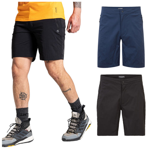 A man stands wearing black shorts with a yellow shirt and hiking shoes displaying a tattoo on his leg while two pairs of shorts are displayed next to him in blue and black.