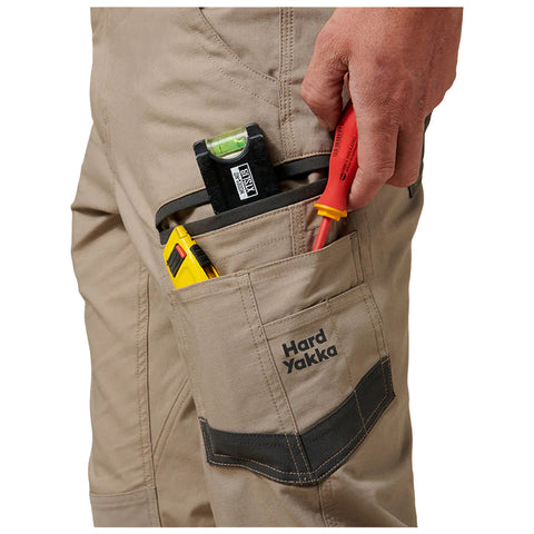 A hand holds a red screwdriver while other tools are stored in a beige pocket of work pants labeled Hard Yakka. The setting suggests a practical work environment.