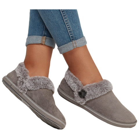 Gray slippers with a fur-lined collar are worn on feet with rolled-up denim jeans. The slippers feature a button detail and have a soft texture, suitable for casual indoor wear.