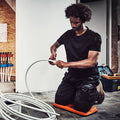 FENTO Board - Ergonomic A man kneels on a flat surface while cutting a white hose with a tool surrounded by coiled hoses in a workshop with various tools and pipes in the background.