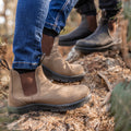 Brown and black boots are positioned on a textured log in a forested area with soft earth and foliage, suggesting outdoor activity or exploration.