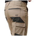 Beige work pants feature multiple pockets and a reinforced design. They are worn by a person facing slightly away, highlighting the brand label and contrasting patches on the fabric.