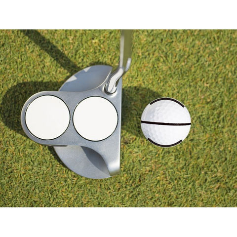 A golf putter with two white circular faces is positioned against a golf ball on green grass. The ball is marked with a black line around its middle, indicating its orientation.