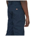 A person stands wearing dark blue work pants with visible pockets and a brand logo on the back waistband while their hand rests at their side in a neutral setting.