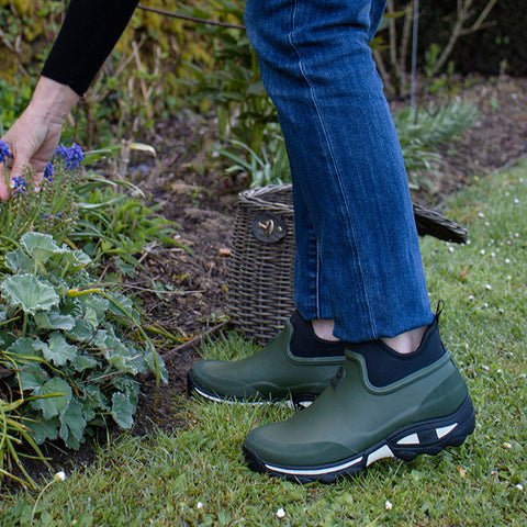 A person wearing green gardening shoes kneels to pick purple flowers from a garden bed while surrounded by lush green plants and a woven basket in the background.