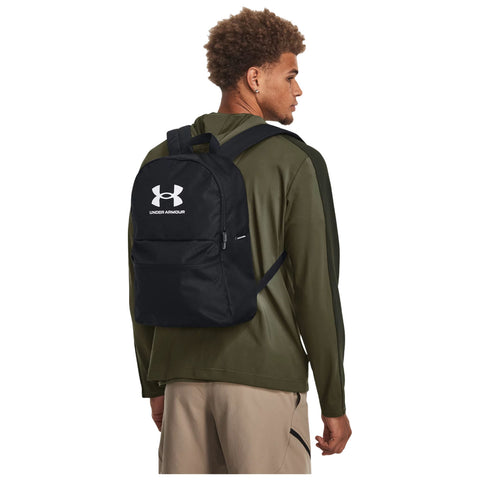 A young man wears a black Under Armour backpack while looking over his shoulder in a casual outdoor setting, dressed in a long-sleeve green top and light-colored pants.