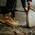 A pair of brown hiking boots stands on gravel while a hand lifts a canoe, suggesting preparation for an outdoor activity near a body of water.