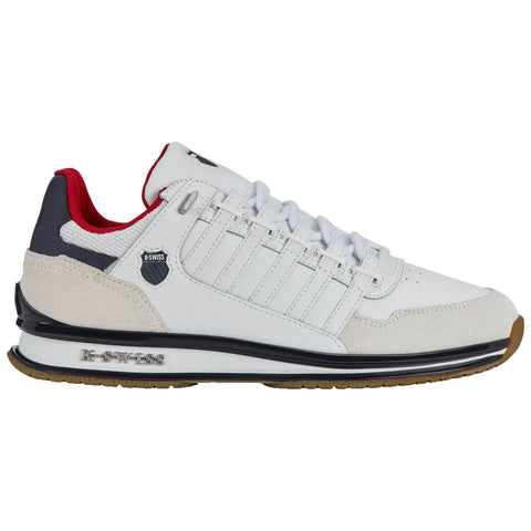 A white athletic shoe with red and navy accents features a lace-up design and suede details along the sides and toe. It is set against a neutral background.