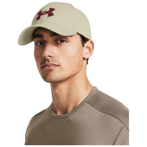 A young man wearing a beige baseball cap with a burgundy logo and a fitted brown shirt is looking slightly to the side against a plain background.