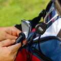 A hand holds a club cleaner brush against a golf club face while a golf bag is nearby on green grass, showcasing the maintenance of golfing equipment in an outdoor setting.