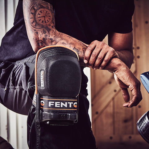 A person's arm covered in intricate tattoos rests on a knee pad labeled FENTO while sitting near a wooden background, indicating a work or construction setting.