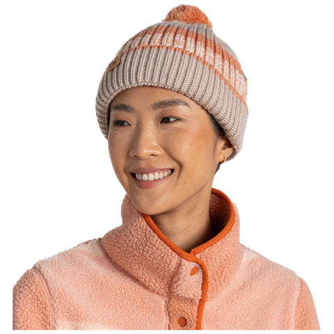 A woman with a warm smile wears a knitted beige and orange striped beanie topped with a pom-pom paired with a light pink fleece jacket featuring an orange collar in a neutral background.