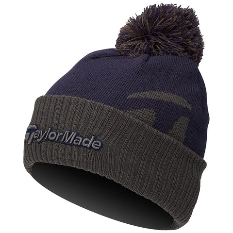 A beanie hat is displayed with a navy blue upper section and a dark green ribbed band at the bottom featuring the text TaylorMade stitched in navy. The hat has a fluffy pom-pom on top.