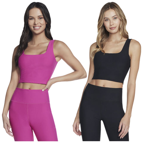 Two women stand side by side wearing matching athletic outfits. One wears a pink crop top and leggings while the other wears a black crop top and leggings, both smiling.