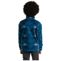 A child with curly hair stands with their back to the viewer wearing a blue patterned fleece jacket and dark pants in a neutral, uncluttered space.