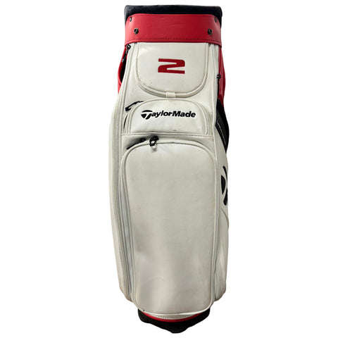 A golf bag stands upright with a white body and red top The bag features the number 2 and the TaylorMade logo along with multiple zippered compartments for storing golf equipment