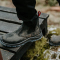 A black boot with a textured surface steps onto a wooden platform surrounded by mossy ground, suggesting outdoor activity and rugged terrain.