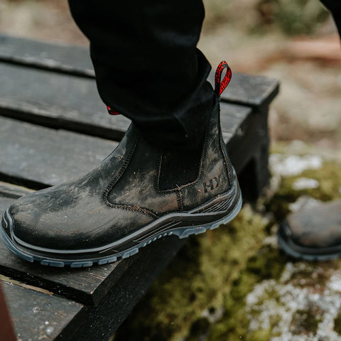 A black boot with a textured surface steps onto a wooden platform surrounded by mossy ground, suggesting outdoor activity and rugged terrain.