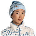 A woman wears a light blue knitted beanie with a pom-pom and stripes while dressed in a cozy white fleece jacket featuring a blue spotted pattern, creating a warm and stylish appearance.