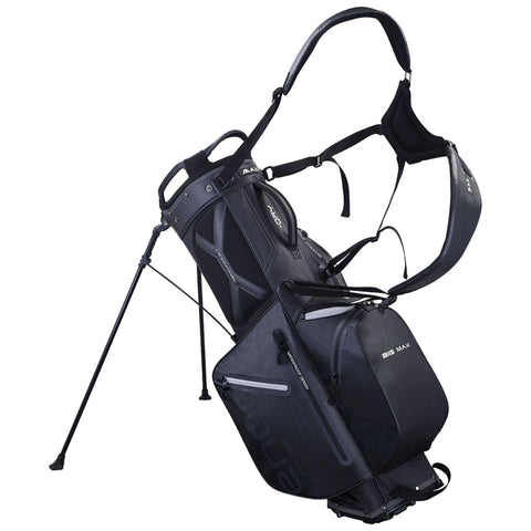 A black golf bag stands upright on three legs featuring a padded shoulder strap and multiple zippered compartments for storage in an outdoor environment.