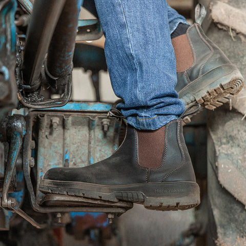 A booted foot rests on a metal step of a tractor with a textured surface while wearing blue jeans in a rustic agricultural setting surrounded by machinery and natural elements.