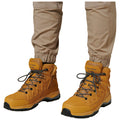 Boots with a rugged design are worn by a person standing. The boots feature a tan color with dark soles and contrasting laces, set against light-colored, tapered pants.