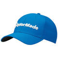 A blue baseball cap displays the logo TaylorMade prominently on the front in white embroidery resting on a flat surface with a simple textured background.