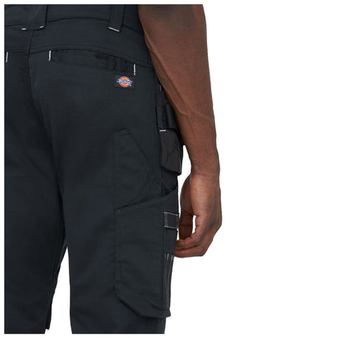 A pair of black work pants is shown with a hand holding onto the waist area highlighting multiple pockets and a brand logo on the back in a neutral background.