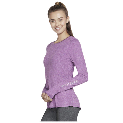 A woman wears a long-sleeved purple athletic top while smiling and posing with her hands clasped at her waist against a plain white background. The top has "SKECHERS" displayed on the sleeve.