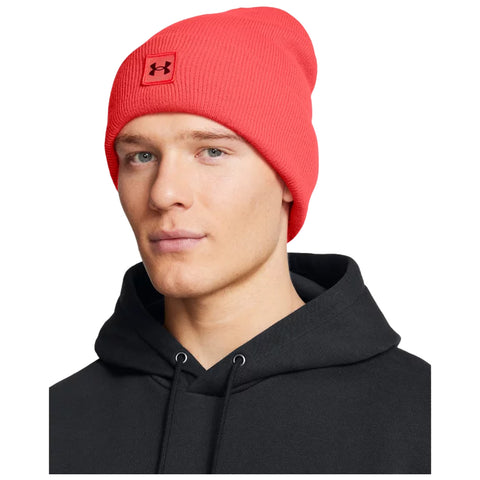 A person wears a bright red beanie with a logo while looking directly at the viewer against a plain background, dressed in a black hooded sweatshirt.