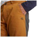 A close-up of yellow-brown pants with a dark side panel and a logo on the thigh a hand is positioned in the pocket against a neutral background