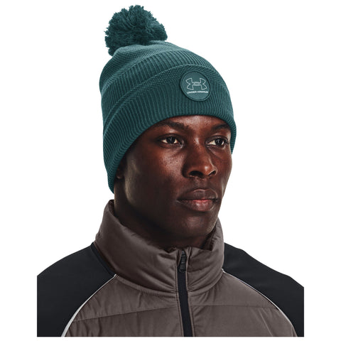 A man wears a teal knit beanie with a pom-pom while dressed in a brown jacket with a high collar in a neutral setting focused on his expression and attire.