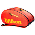 A bright red sports bag with the name Wilson prominently displayed in yellow is positioned horizontally featuring a zippered pocket and dual carrying handles for transport.