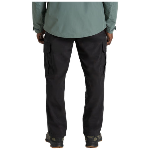 A person stands with their back facing the viewer wearing a green long-sleeve shirt and black cargo pants with side pockets and sturdy footwear. The setting is neutral and simple.