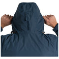 A person wearing a blue hooded jacket pulls the hood closer using both hands while standing against a plain white background. The jacket features an adjustment toggle at the neck.