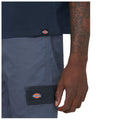 A hand reaches toward a pocket on gray work pants featuring a Dickies logo while wearing a dark blue shirt with a smaller logo on the hem, showcasing a casual workwear style.