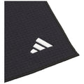 A black textile object displays a three-stripe logo in white at one corner while resting flat on a surface, exhibiting a textured pattern throughout its body.