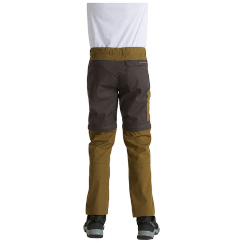 A child is seen from behind wearing two-tone pants with a combination of dark and light brown colors while standing on a flat surface with a white background.